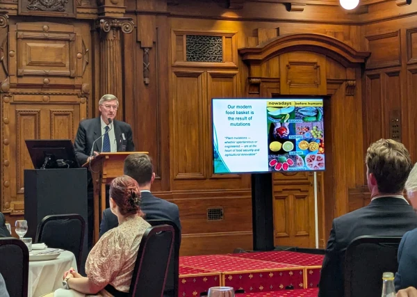 Grey-haired Dr Caradus leans forward at the podium next to a slide showing colourful fruit and vegetables. Photo taken through dinging guests in the foreground.