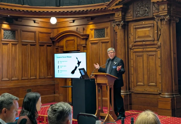 Prof Dearden gesticulates behind a podium in a wood paneled room with red carpet. He stands next to a large screen with a slide showing a silhouette of New Zealand and the heads of seated MPs are visible in the foreground.