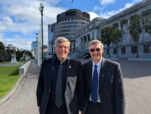 Two white men in suits stand in the sun in front of the beehive