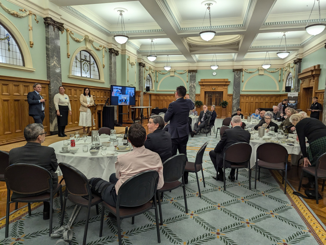 Prof Richard Easther, Dr Pauline Harris, and host Dr Parmjeet Parmar MP listen to a question from an MP Prof Richard Easther, Dr Pauline Harris, and host Dr Parmjeet Parmar MP listen to a question from a male MP, who stands at a round table, amid seated guests, with his back to the camera